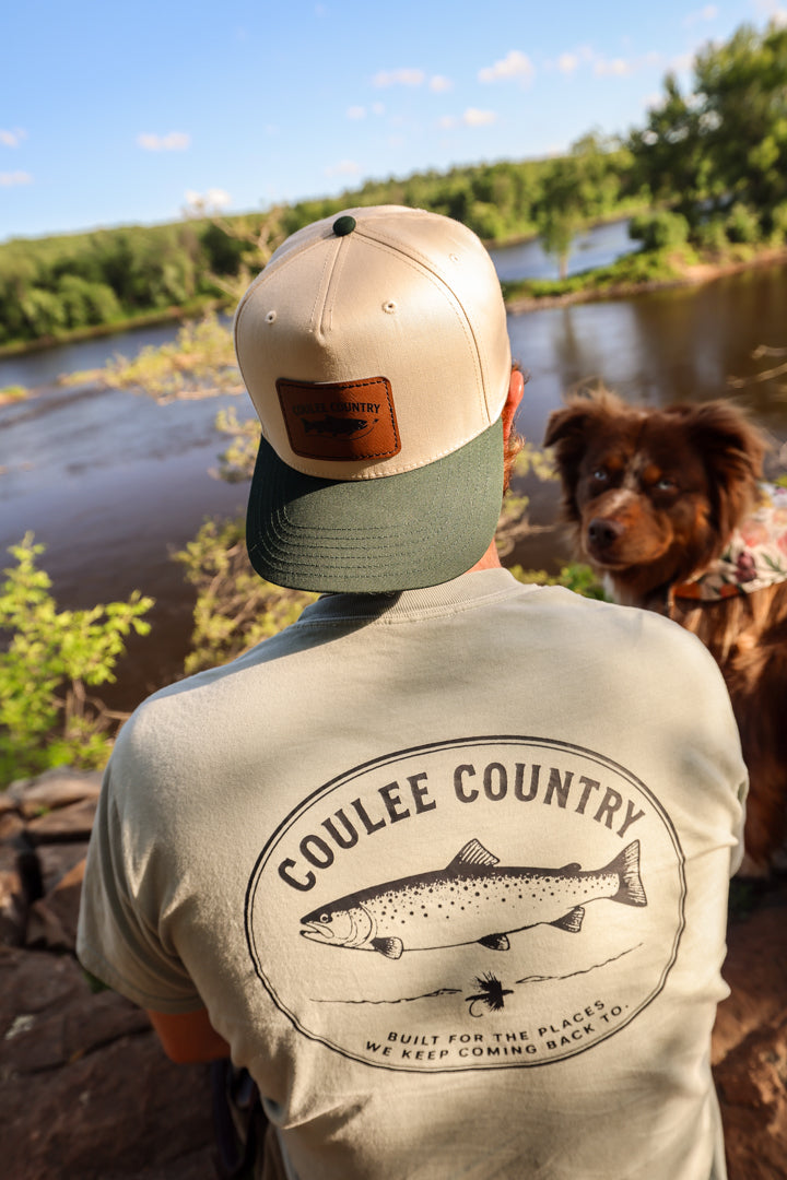 Man wearing the Coulee Country River Run Pocket Tee in sage with back trout graphic, sitting above a river with his dog
