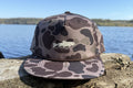 Coulee Country Flyway Rope Hat in bark duck camo with cream trout logo, on a driftwood log by the river