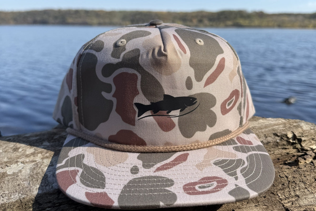 Coulee Country Flyway Rope Hat in harvest duck camo with black trout logo, on a driftwood log by the river