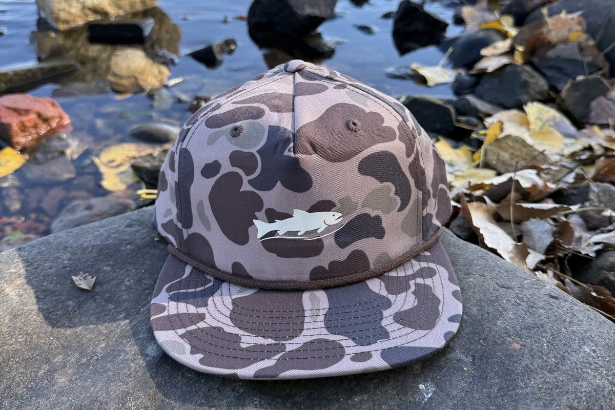 Coulee Country Flyway Rope Hat in bark duck camo with cream trout logo, on a riverside rock with fall leaves